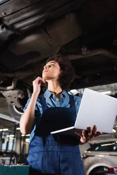 Joven afroamericano mecánico inspeccionar coche levantado con portátil en la mano en el garaje - foto de stock