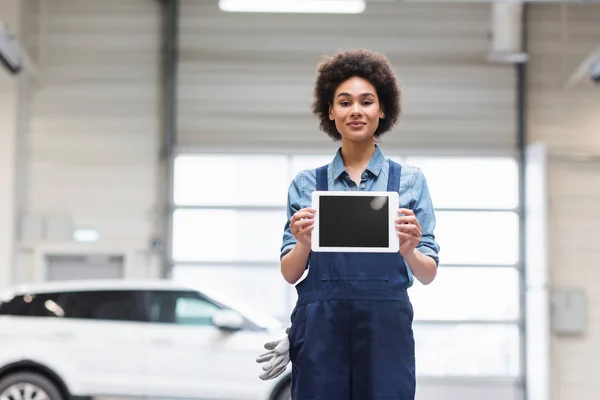 Smiling young african american mechanic showing digital tablet with blank screen and looking at camera in garage — Stock Photo