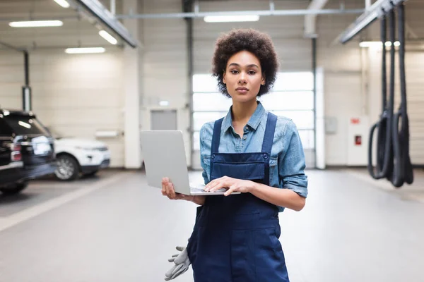 Ernsthafter junger afrikanisch-amerikanischer Mechaniker steht mit Laptop in der Garage und schaut in die Kamera — Stockfoto