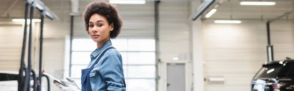 Confident young african american mechanic standing with digital tablet and looking at camera in garage, banner — Stock Photo