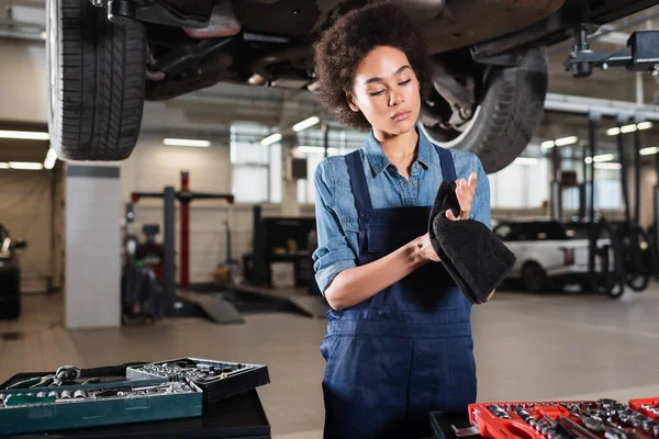 Fokussierter junger afrikanisch-amerikanischer Mechaniker steht unter Auto und trocknet Hände mit Handtuch in Garage — Stockfoto