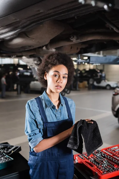 Jovem afro-americano mecânico em pé debaixo do carro e secar as mãos com toalha na garagem — Fotografia de Stock