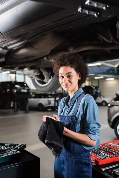 Lächelnde junge afrikanisch-amerikanische Mechaniker in Overalls trocknen Hände mit Handtuch in der Garage — Stockfoto