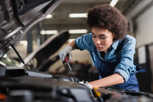 Young african american mechanic repairing car motor with screwdriver in garage — Stock Photo