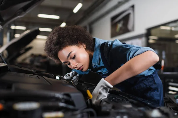 Young african american mechanic fixing motor in car with open hood in garage — Stock Photo