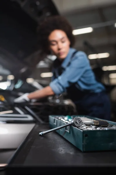 Nahaufnahme von Schraubenschlüssel in Werkzeugkiste mit verschwommenem afrikanisch-amerikanischem Mechaniker bei der Arbeit mit Auto in Garage — Stockfoto