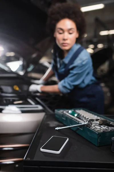 Vista da vicino di cellulare e chiave inglese in cassetta degli attrezzi con donna afroamericana offuscata che lavora in garage — Foto stock