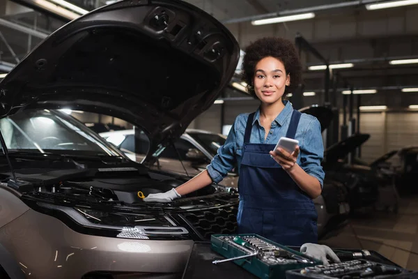 Lächelnder junger afrikanisch-amerikanischer Mechaniker steht mit Handy neben Auto mit offener Motorhaube in Garage — Stockfoto