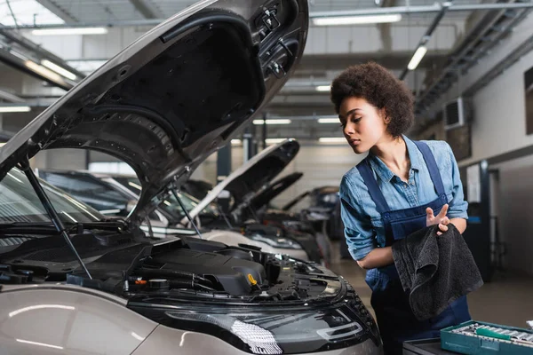 Giovane afro americano meccanico asciugatura mani con asciugamano vicino auto con cappuccio aperto in garage — Foto stock