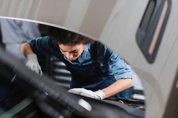 Young african american mechanic in overalls holding pliers and working with car hood in garage — Stock Photo