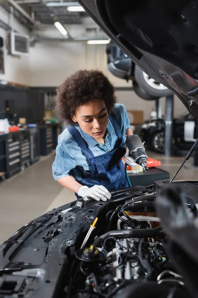 Young african american mechanic holding electric screwdriver and looking at motor of car with open hood in garage — Stock Photo
