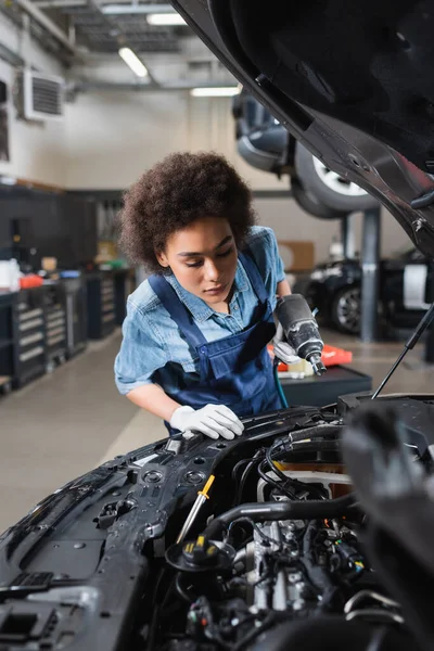 Giovane meccanico afroamericano che tiene in mano un cacciavite elettrico e guarda il motore di un'auto con cappuccio aperto in garage — Foto stock