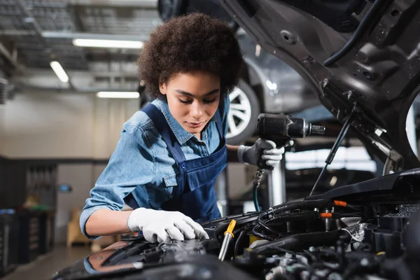 Joven mecánico afroamericano en overoles sosteniendo destornillador eléctrico y trabajando con motor de coche en garaje - foto de stock