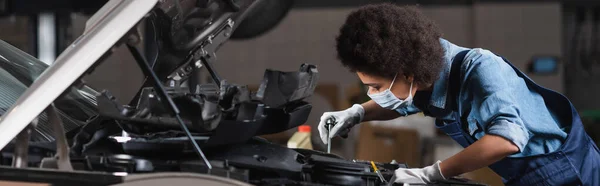 Joven mecánico afroamericano en máscara protectora que sostiene la llave inglesa y que trabaja con el motor del coche en el garaje, bandera - foto de stock