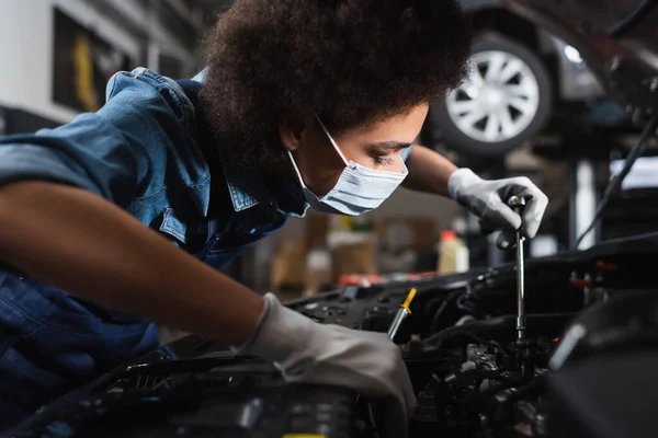 Jovem afro-americano mecânico em máscara protetora trabalhando com motor de carro na garagem — Fotografia de Stock