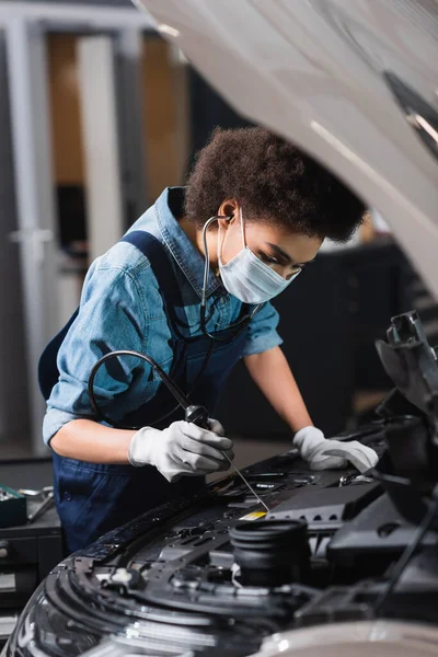Young african american mechanic in protective mask and stethoscope working with car motor in garage — Stock Photo