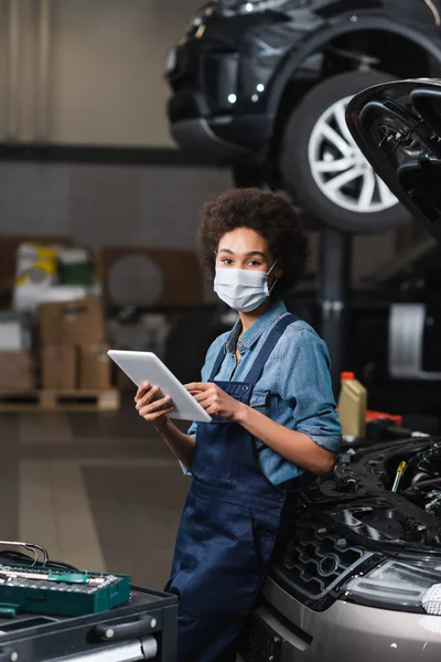 Joven afroamericano mecánico en máscara protectora celebración tableta digital cerca de coche en garaje — Stock Photo