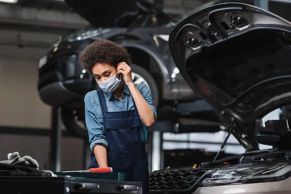 Jovem afro-americano mecânico ajustando máscara protetora enquanto trabalhava perto de carro na garagem — Fotografia de Stock