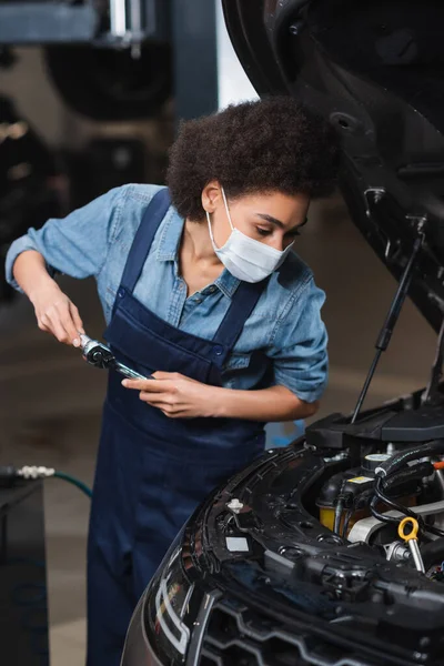 Focado jovem afro-americano mecânico em máscara protetora segurando chave inglesa e trabalhando com motor de carro na garagem — Fotografia de Stock