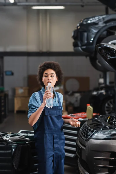 Joven africano americano mecánico beber agua de botella cerca de coche en garaje - foto de stock