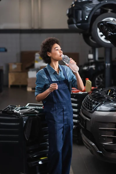 Joven afroamericano mecánico en overoles beber agua de botella cerca de coche en garaje - foto de stock