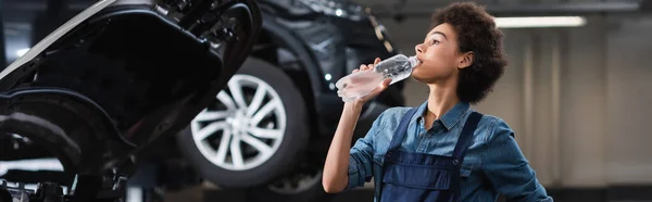 Joven afroamericano mecánico en overoles beber agua de botella cerca de coche en garaje, pancarta - foto de stock