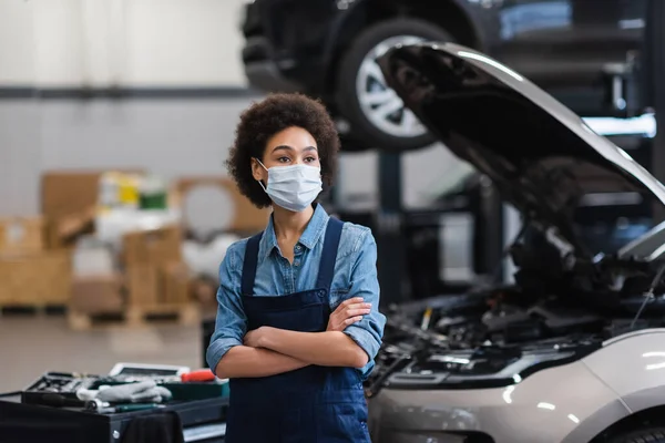 Pensativo jovem afro-americano mecânico em máscara protetora de pé com braços cruzados perto do carro na garagem — Fotografia de Stock