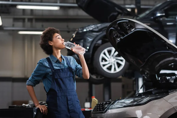 Junger afrikanisch-amerikanischer Mechaniker in Overalls trinkt Wasser aus Plastikflasche neben Auto in Garage — Stockfoto