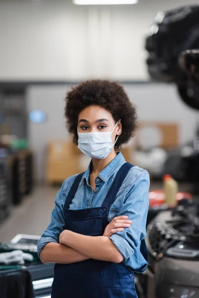 Young african american mechanic in protective mask standing with crossed arms near car in garage — Stock Photo