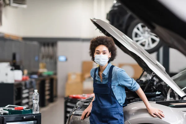 Joven afroamericano mecánico en máscara protectora de pie cerca de coche en garaje - foto de stock