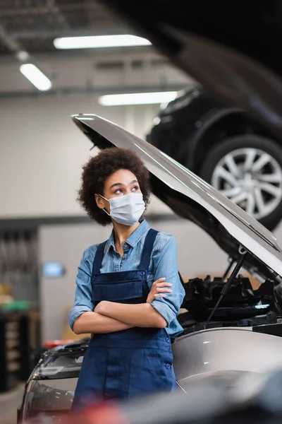 Jovem afro-americano mecânico em máscara protetora de pé com braços cruzados perto de carro com capuz aberto na garagem — Fotografia de Stock