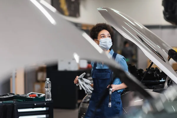 Jeune mécanicien afro-américain en masque de protection tenant l'équipement près des voitures avec hottes ouvertes dans le garage — Photo de stock