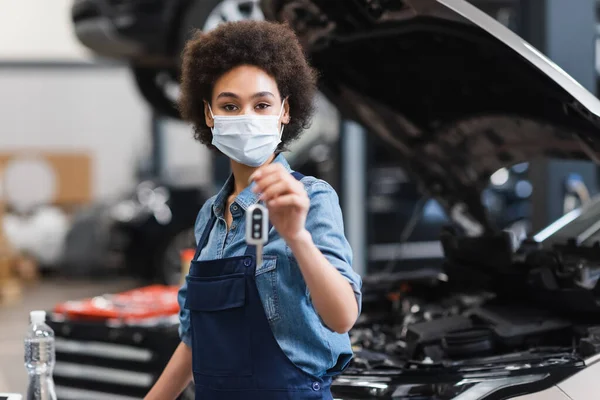 Young african american mechanic in protective mask holding blurred car key in hand in garage — Stock Photo