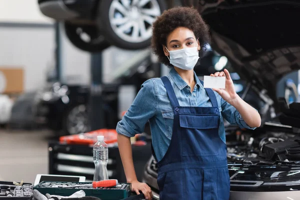 Young african american mechanic in protective mask showing empty card in garage — Stock Photo