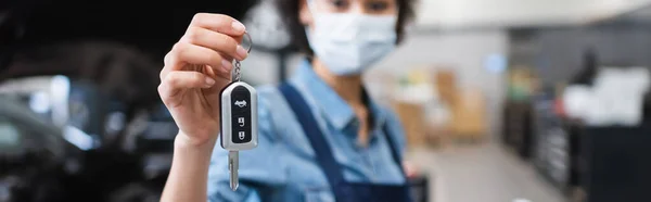 Close up view of car key in hand of blurred young african american mechanic in protective mask in garage, banner — Stock Photo
