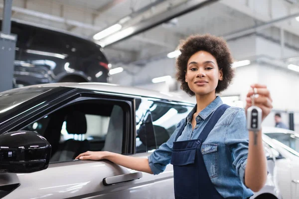 Lächelnder junger afrikanisch-amerikanischer Mechaniker in Overalls, der den Schlüssel neben dem Auto in der Garage hält — Stockfoto