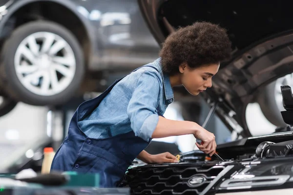 Jovem afro-americano mecânico segurando chave de fenda e motor de fixação de carro na garagem — Fotografia de Stock