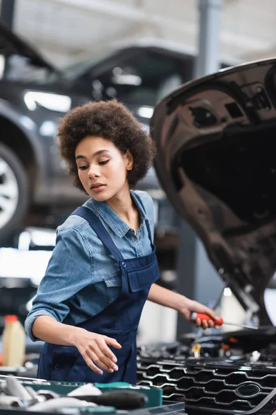 Joven afroamericano mecánico sosteniendo destornillador y trabajando con motor de coche en garaje — Stock Photo