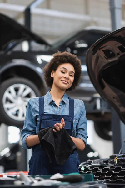 Sorridente giovane meccanico afroamericano in tuta asciugandosi le mani con asciugamano nero in garage — Foto stock