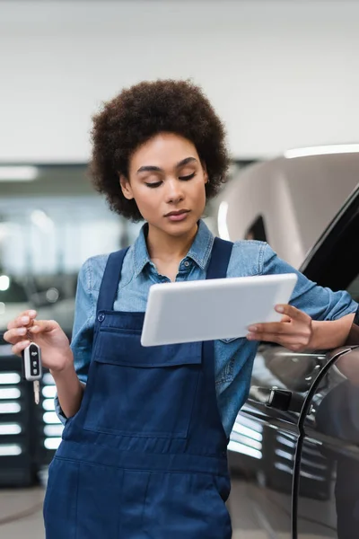 Young african american mechanic holding car key and looking at digital tablet in garage — Stock Photo