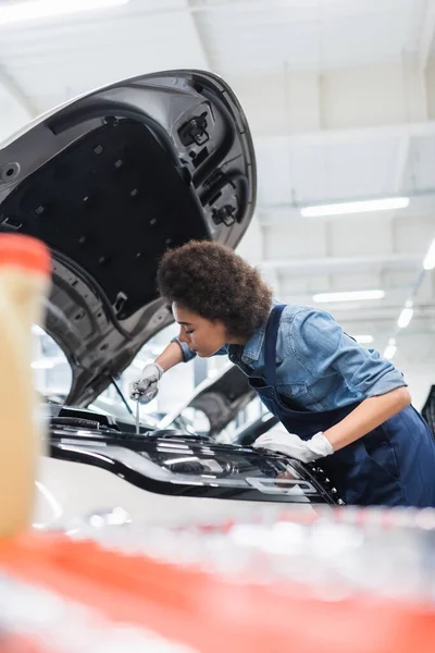Young african american mechanic fixing motor in car with open hood in garage — Stock Photo