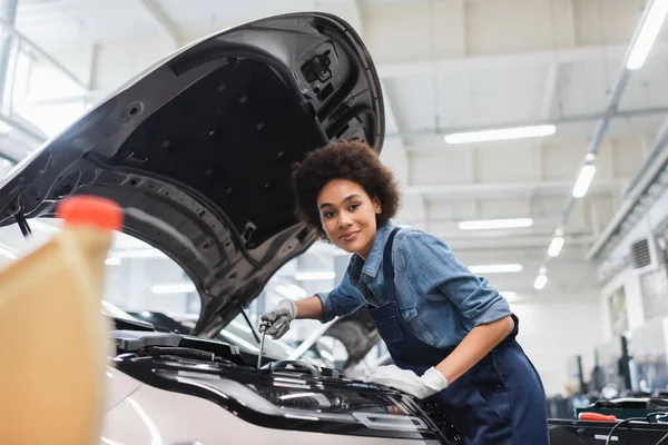 Smiling young african american mechanic fixing motor in car with open hood in garage — Stock Photo