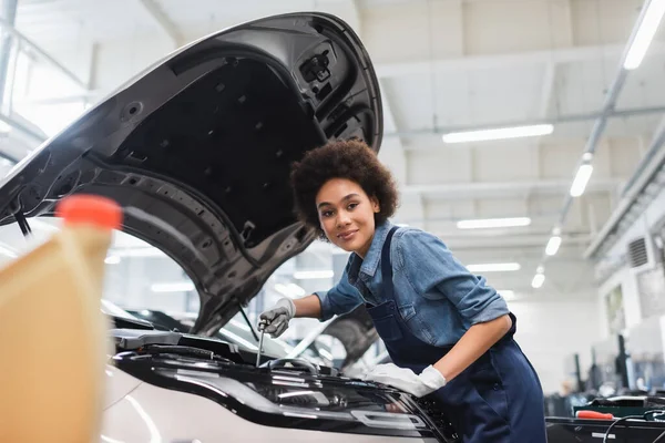 Sorridente giovane afroamericano meccanico di fissaggio motore in auto con cappa aperta in garage — Foto stock