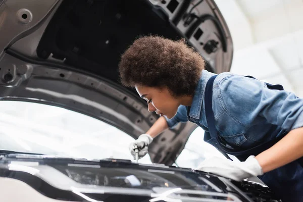 Young african american mechanic fixing motor in car with open hood in garage — Stock Photo