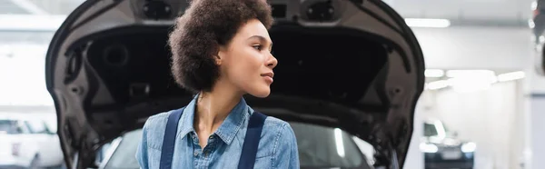 Joven afroamericano mecánico de pie cerca de capucha abierta y mirando hacia otro lado en el servicio de reparación de automóviles, pancarta - foto de stock