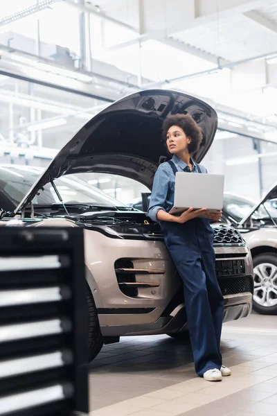 Giovane meccanico afroamericano in piedi con computer portatile vicino alle auto con cappuccio aperto nel servizio di riparazione auto — Foto stock