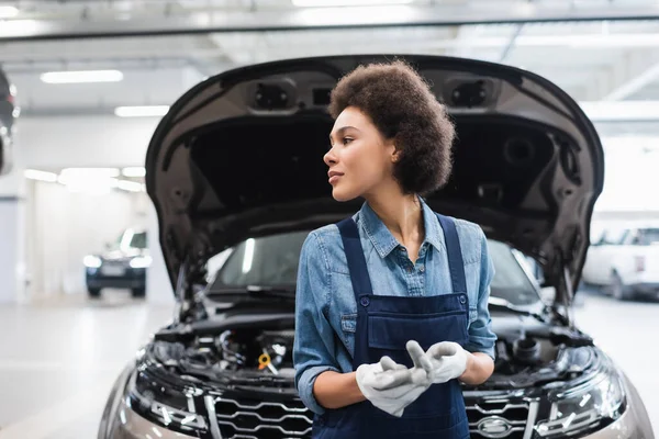 Joven afroamericano mecánico de pie cerca de coche con capucha abierta y mirando hacia otro lado en el servicio de reparación de automóviles — Stock Photo