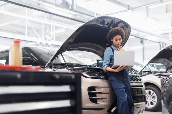 Young african american mechanic holding laptop near car with open hood in auto repair service — Stock Photo