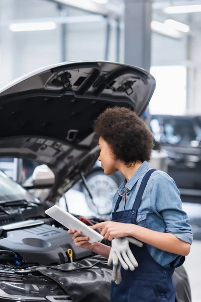 Junger afrikanisch-amerikanischer Mechaniker hält digitales Tablet in der Hand und betrachtet Auto mit offener Motorhaube im Autoreparaturservice — Stockfoto