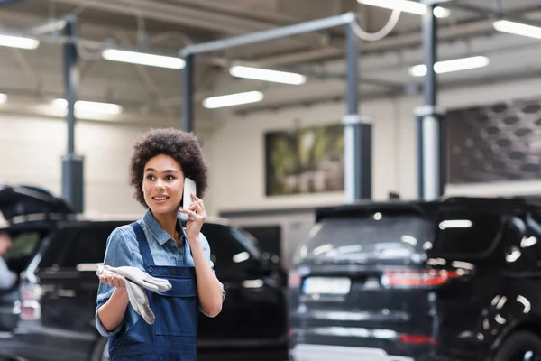 Worried young african american mechanic in overalls speaking on smartphone in auto repair service — Stock Photo